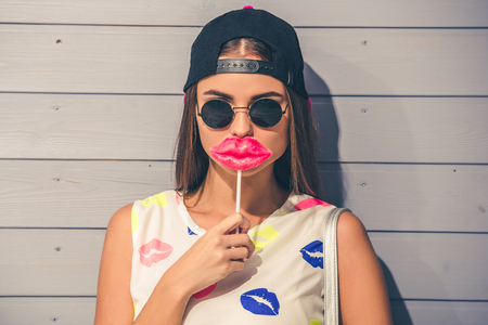 Stylish teenage girl in sun glasses and cap is holding a lips-lollipop and looking at camera, on gray backgroundの写真素材
