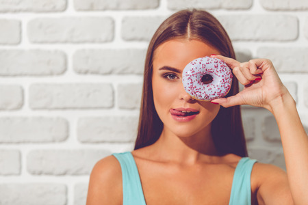 Portrait of funny girl holding a doughnut on eye, looking at camera and licking lips, on white brick wall backgroundの写真素材