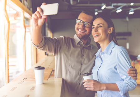 Handsome businessman and beautiful business lady are doing selfie using a smartphone, holding cups of coffee and smiling while co-workingの写真素材