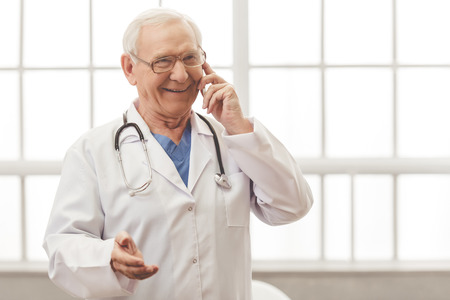 Handsome old doctor in white medical coat and eyeglasses is talking on the mobile phone and smiling while standing against windowの写真素材