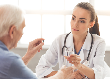 Beautiful female doctor in white medical coat is consulting her old patient and giving him glass of water. Man is holding a pillの写真素材