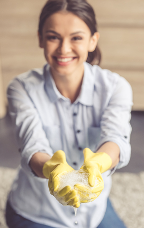 Beautiful young woman is holding soapsuds, looking at camera and smiling while cleaning the house. Hands in focusの写真素材