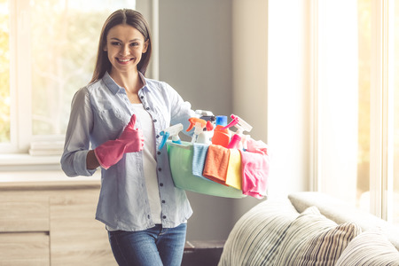 Beautiful woman is holding a basin with detergents and rags, showing Ok sign, looking at camera and smiling ready to clean her houseの写真素材