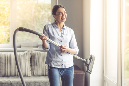 Beautiful young woman in earphones is imitating playing guitar using a vacuum cleaner and smiling while cleaning her houseの写真素材