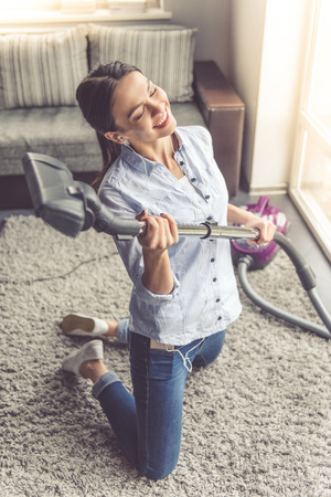 Beautiful young woman in earphones is imitating playing guitar using a vacuum cleaner and smiling while cleaning her houseの写真素材