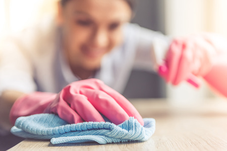 Woman in protective gloves is smiling and wiping dust using a spray and a duster while cleaning her...