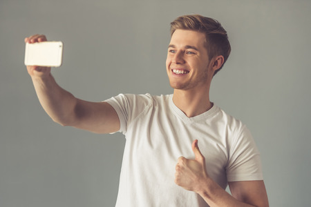 Handsome guy in white t-shirt is doing selfie using a smart phone, showing Ok sign and smiling, on gray backgroundの写真素材