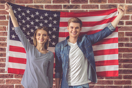 Beautiful young couple is holding American flag, looking at camera and smiling, on brick wall backgroundの写真素材