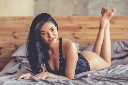 Sexy young woman in black lingerie is looking sensually at camera while lying on bedの写真素材