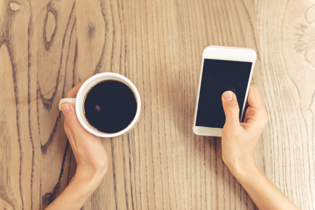 Cropped image of beautiful young woman using a smartphone and holding a cup of coffeeの写真素材