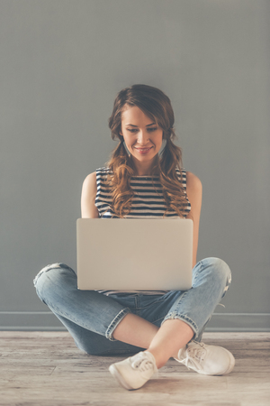 Stylish young woman is using a laptop and smiling while sitting on the floor, on gray backgroundの写真素材