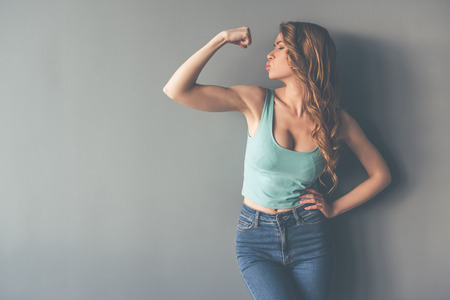 Beautiful stylish young woman is showing her muscles and sending kiss, on gray background