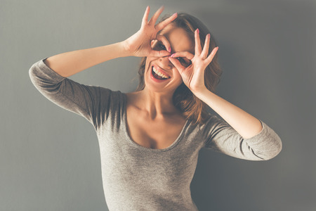 Beautiful stylish young woman is showing glasses, looking at camera and smiling, on gray backgroundの写真素材
