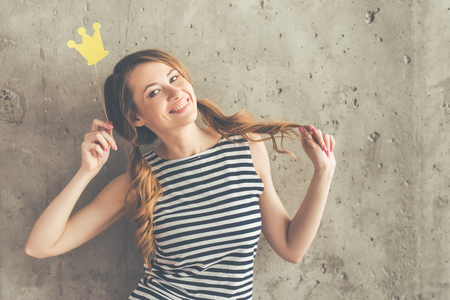 Stylish young woman in sun glasses is holding a paper crown on stick, looking at camera and smiling, on beton wall backgroundの写真素材