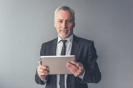 Handsome mature businessman in formal suit is using a digital tablet, looking at camera and smiling, on gray backgroundの写真素材