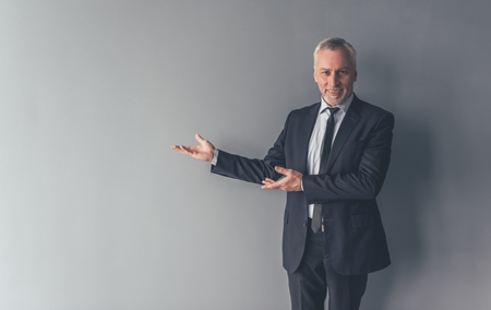 Handsome mature businessman in formal suit is pointing away, looking at camera and smiling, on gray backgroundの写真素材