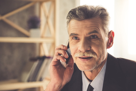 Handsome mature businessman in classic clothes is talking on the mobile phone and smiling while working in officeの写真素材