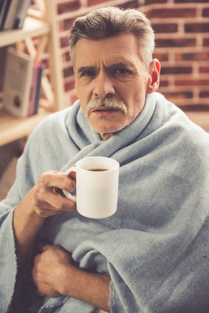 Handsome ill mature man is drinking hot tea while sitting wrapped in blanket at homeの写真素材
