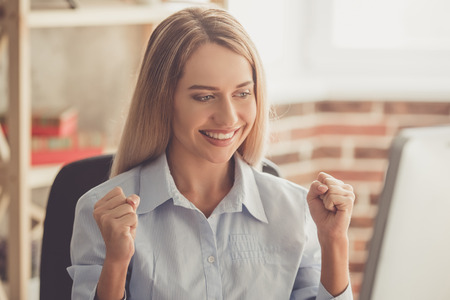 Beautiful business lady in formal shirt is raising hands in fists and smiling while working with a computer in the officeの写真素材