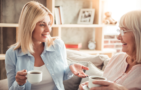 Beautiful mature mother and her adult daughter are drinking coffee, talking and smiling while sitting on couch at homeの写真素材