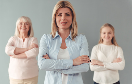 Three generations of women. Beautiful granny, mother and daughter are looking at camera and smiling, standing with crossed arms on gray backgroundの写真素材