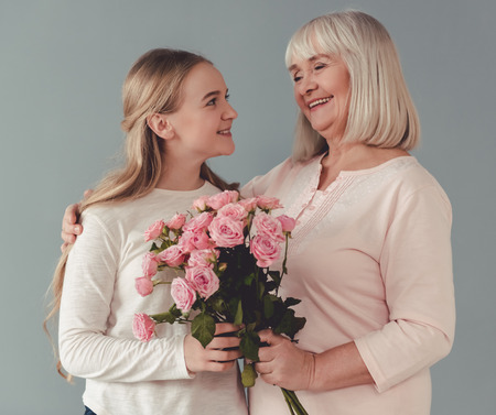Cute teenage girl is giving flowers to her beautiful granny, on gray background. Both are smiling