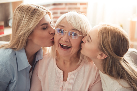 Three generations of women. Beautiful woman and teenage girl are kissing their granny while sitting on couch at homeの写真素材