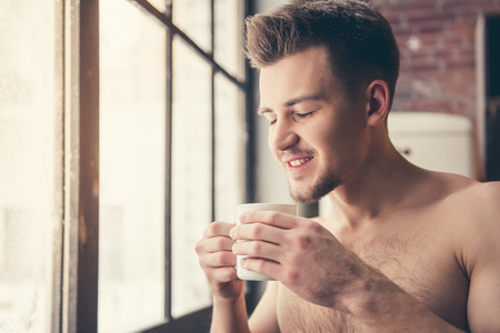 Sexy muscular guy with bare torso is holding a cup and smiling while standing in the kitchenの写真素材