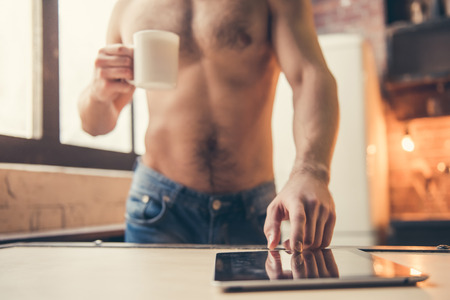 Cropped image of sexy muscular guy with bare torso using a digital tablet and holding a cup while standing in the kitchenの写真素材