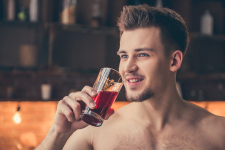 Sexy muscular guy with bare torso is drinking juice and smiling while standing in the kitchenの写真素材