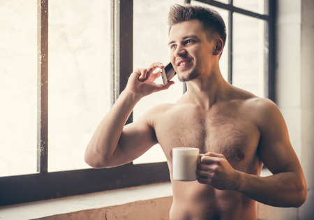 Sexy muscular guy with bare torso is holding a cup, talking on the mobile phone and smiling while standing in the kitchenの写真素材