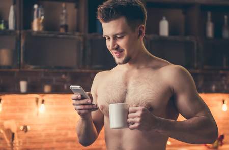 Sexy muscular guy with bare torso is using a smartphone, holding a cup and smiling while standing in the kitchenの写真素材