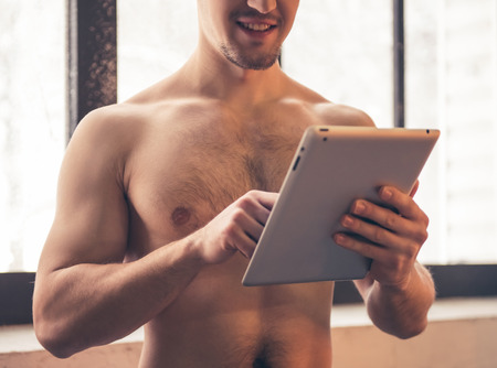 Sexy muscular guy with bare torso is using a digital tablet and smiling while standing in the kitchenの写真素材