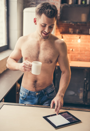Sexy muscular guy with bare torso is using a digital tablet, drinking coffee and smiling while standing in the kitchenの写真素材