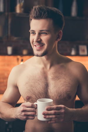 Sexy muscular guy with bare torso is holding a cup and smiling while standing in the kitchenの写真素材