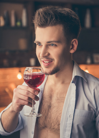Sexy guy in unbuttoned shirt is drinking wine and smiling while standing in the kitchenの写真素材