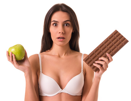Beautiful slim girl in white bra is holding an apple and a bar of chocolate, looking at camera and showing confusion, isolated on whiteの写真素材