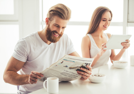 Beautiful young couple is having a breakfast together at home. Girl is using a digital tablet, guy is reading a newspaperの写真素材