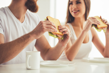 Beautiful young couple is eating sandwiches, looking at each other and smiling while having a breakfast at homeの写真素材