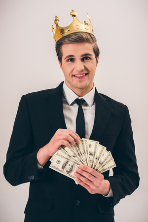 Handsome young man in suit and crown is holding money, on gray backgroundの写真素材