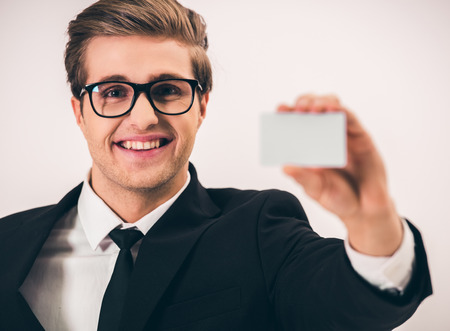 Handsome young businessman in suit and eyeglasses is holding a card, looking at camera and smiling, on gray backgroundの写真素材