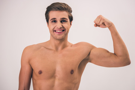 Portrait of handsome young naked man showing biceps, looking at camera and smiling, on gray backgroundの写真素材
