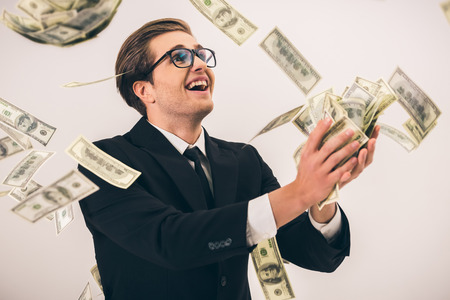 Handsome young businessman in suit and glasses is catching cash and smiling, on gray backgroundの写真素材