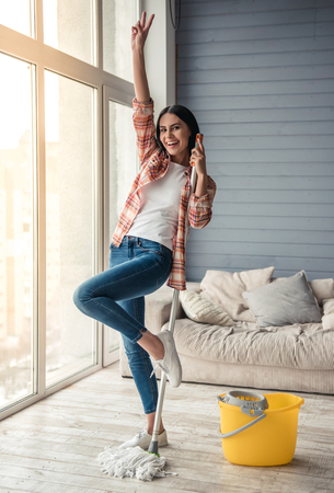 Beautiful young woman is dancing and smiling while cleaning floor at home using a mopの写真素材