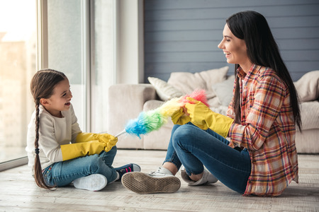 Beautiful young woman and her little daughter are sitting on the floor and smiling while cleaning...