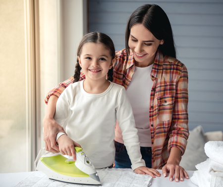 Beautiful young woman and her little daughter are smiling while ironing linenの写真素材