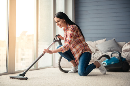 Beautiful young woman is smiling and using a vacuum cleaner while cleaning floor at homeの写真素材