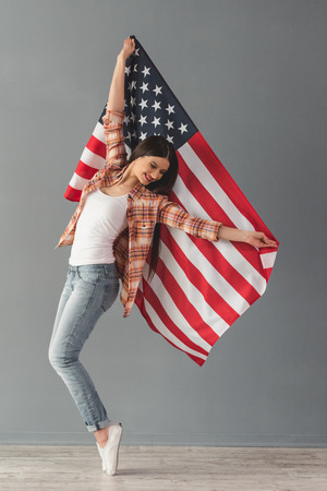 Cheerful young woman in casual clothes is holding the American flag and smiling while standing on her toes, on gray backgroundの写真素材
