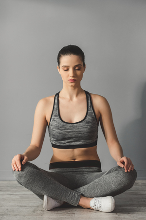 Attractive young woman in sportswear is sitting in lotus position while doing yoga, on gray backgroundの写真素材