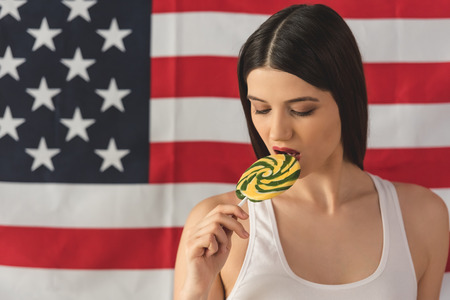Cheerful young woman is tasting a lollipop, on American flag backgroundの写真素材